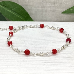 May include: A delicate silver chain bracelet with red gemstone beads. The bracelet has a lobster clasp closure.
