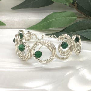 May include: A silver wire bracelet with green gemstone detailing. The bracelet has a series of looped wire designs, each holding a small, round, green gemstone. The bracelet is shown on a white surface with green foliage in the background.