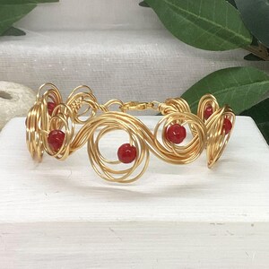 May include: A gold wire bracelet with red beads. The bracelet is made of wire that is twisted into a spiral pattern. The beads are evenly spaced along the bracelet.