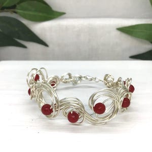 May include: A silver wire bracelet with red beads. The bracelet has a series of swirling wire designs, each with a small, round, red bead. The clasp is a simple silver hook and eye closure. The bracelet is displayed on a white surface.