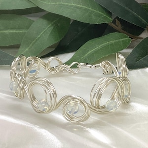 May include: A silver-toned wire bracelet featuring a series of interconnected circular wire coils. Each coil secures a small, translucent, iridescent bead. The bracelet is fastened with a simple metal clasp, creating a continuous, linked design.