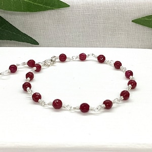 May include: A bracelet with deep red, faceted beads, strung on silver-coloured wire. The bracelet has a lobster clasp and an extension chain for adjustment. The beads are evenly spaced, creating a simple and elegant design. The bracelet's length is adjustable.