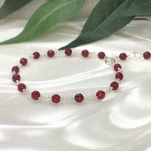 May include: A delicate bracelet featuring small, faceted, deep red beads linked together by thin silver-toned wire. The design includes a short extender chain with a small lobster claw clasp, creating a continuous, lightweight circular chain structure.