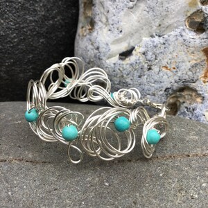 May include: A silver wire bracelet with turquoise beads. The bracelet is made of many small, intertwined loops of wire, creating a delicate and intricate design.