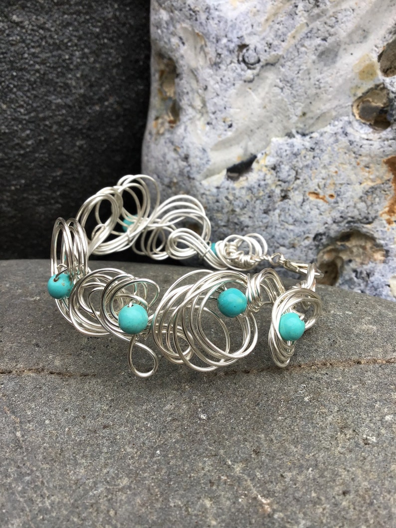 May include: A silver wire bracelet with turquoise beads. The bracelet is made of many small, intertwined loops of wire, creating a delicate and intricate design.