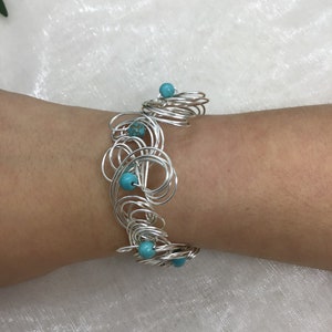May include: A silver wire bracelet with turquoise beads. The bracelet is made of many small circles of wire that are intertwined. The turquoise beads are scattered throughout the bracelet.
