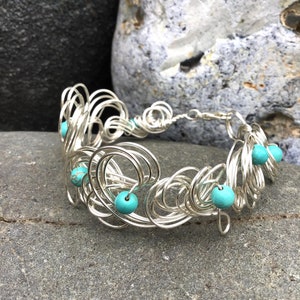 May include: Silver wire bracelet with turquoise beads. The bracelet is made of multiple loops of wire, creating a delicate and intricate design.