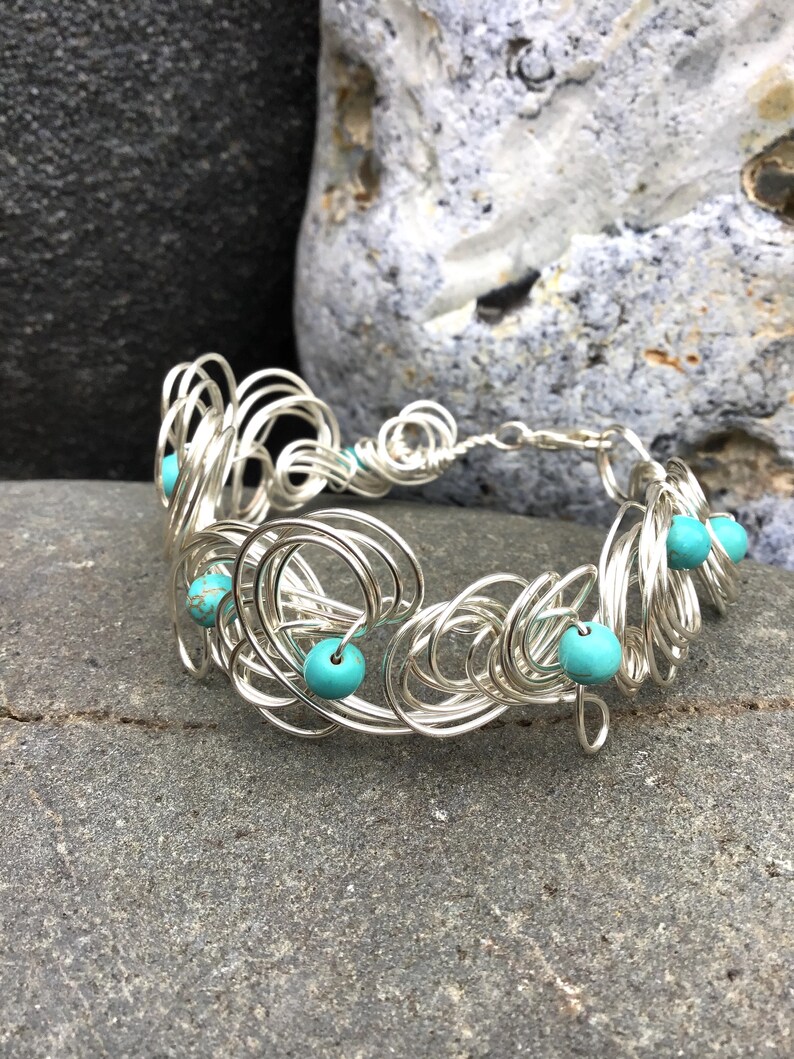 May include: Silver wire bracelet with turquoise beads. The bracelet is made of multiple loops of wire, creating a delicate and intricate design.