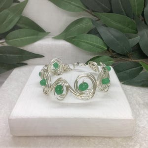 May include: A silver wire bracelet with green glass beads. The bracelet is made of wire that is twisted into a spiral pattern. The beads are evenly spaced along the bracelet.