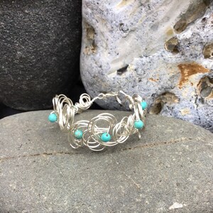 May include: A silver wire bracelet with turquoise beads. The bracelet is made of a series of interconnected wire loops, creating a delicate and intricate design.