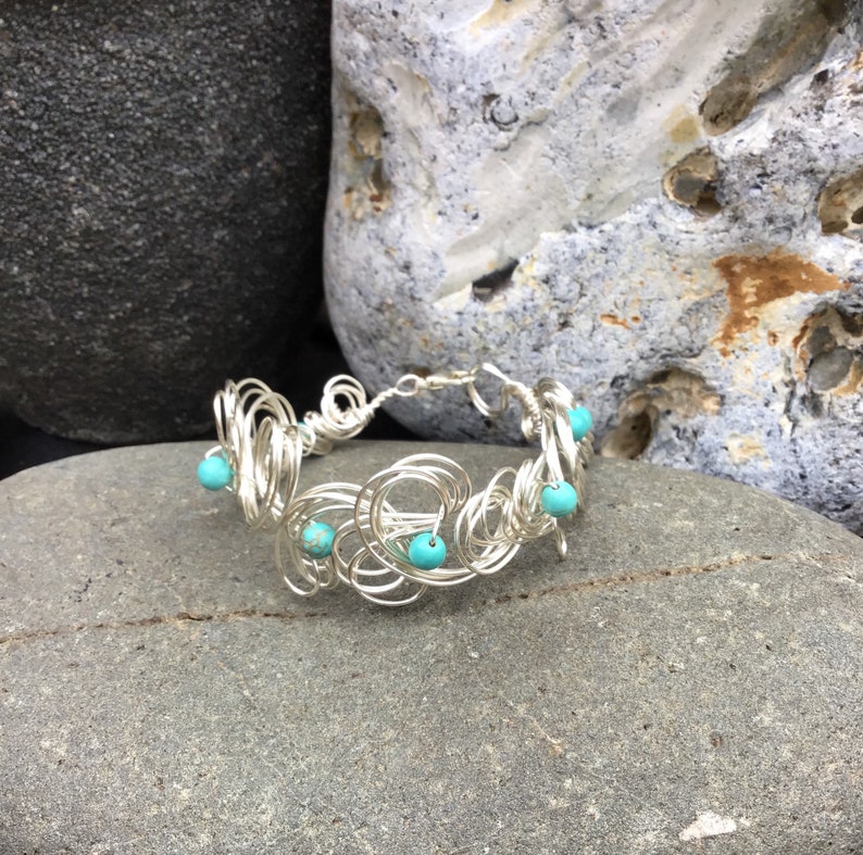 May include: A silver wire bracelet with turquoise beads. The bracelet is made of a series of interconnected wire loops, creating a delicate and intricate design.