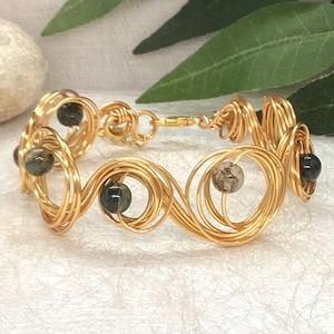 May include: Gold-coloured wire bracelet with a swirling design, featuring small, dark-coloured beads. The bracelet has a clasp closure and is displayed on a white surface, with green foliage in the background. A unique piece of jewellery.