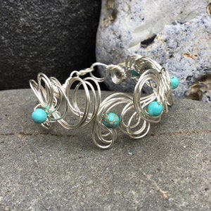 May include: A silver wire bracelet with turquoise beads. The bracelet is made of multiple loops of wire that are intertwined. The turquoise beads are scattered throughout the bracelet.