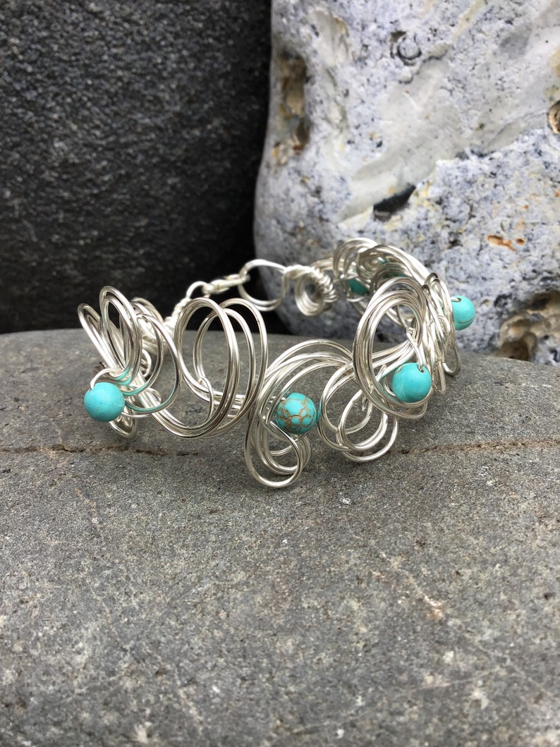 May include: A silver wire bracelet with turquoise beads. The bracelet is made of multiple loops of wire that are intertwined. The turquoise beads are scattered throughout the bracelet.