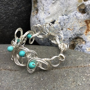 May include: A silver wire bracelet with turquoise beads. The bracelet is made of wire that is twisted and shaped into a delicate design. The beads are evenly spaced along the bracelet.