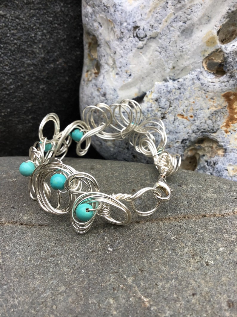 May include: A silver wire bracelet with turquoise beads. The bracelet is made of wire that is twisted and shaped into a delicate design. The beads are evenly spaced along the bracelet.