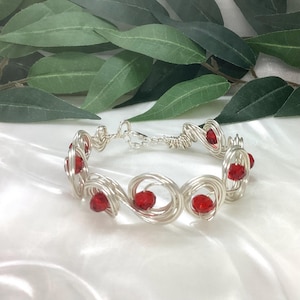 May include: A silver-toned wire-wrapped bracelet featuring multiple circular loops of wire. Each loop contains a single faceted, bright red bead. The bracelet is secured with a small silver-toned metal lobster claw clasp and matching jump ring.