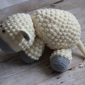 May include: A crocheted white sheep plush toy with gray accents on the face and feet. The sheep is lying on its side with its head turned to the left.