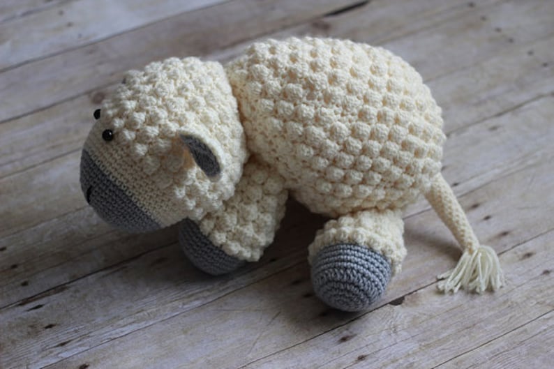 May include: A crocheted white sheep plush toy with gray accents on the face and feet. The sheep is lying on its side with its head turned to the left.