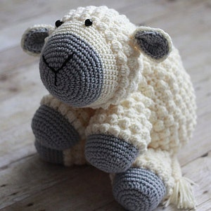 May include: A crocheted white and gray stuffed sheep toy with black eyes. The sheep has a fluffy tail and is sitting on a wooden surface.