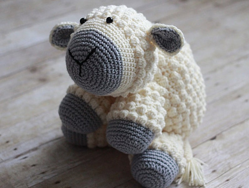 May include: A crocheted white and gray stuffed sheep toy with black eyes. The sheep has a fluffy tail and is sitting on a wooden surface.