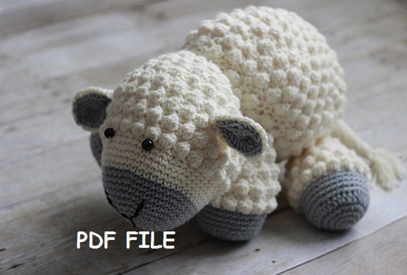 May include: A crocheted white and gray sheep plush toy with black eyes. The sheep has a textured, bumpy surface. The toy is lying on a light wood surface.  PDF FILE.