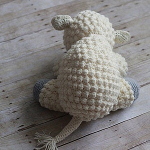May include: A white crocheted stuffed animal shaped like a lamb with gray crocheted feet. The lamb is sitting on a light brown wooden surface.