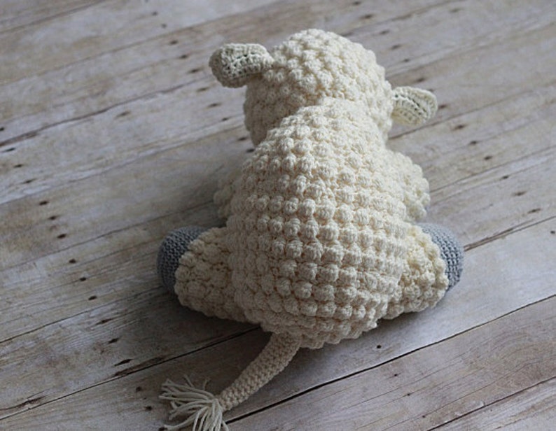 May include: A white crocheted stuffed animal shaped like a lamb with gray crocheted feet. The lamb is sitting on a light brown wooden surface.