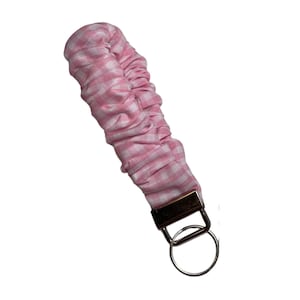 May include: A pink and white gingham fabric wristlet keychain. The fabric is gathered and attached to a gold-coloured metal clasp and a silver-coloured key ring. The wristlet is designed to be worn on the wrist.