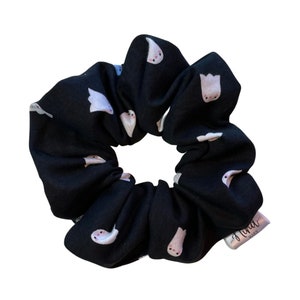 Op de afbeelding: Zwarte scrunchie met een herhaald patroon van witte spookillustraties. De spoken hebben roze wangen en zijn verspreid over de stof. Een klein wit label met de tekst "Luna Marie" is aan de scrunchie bevestigd.