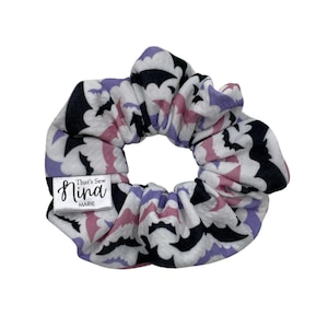 May include: A white scrunchie with a black bat pattern and pink and purple accents. The scrunchie has a label that says "That's Sew Nina MARIE".