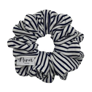 May include: A white and navy blue striped scrunchie. The fabric is gathered and ruched, creating a voluminous effect. A small white tag with the text "Nina MARIE" is attached to the scrunchie.
