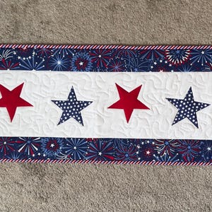 May include: A rectangular quilted table runner with a patriotic theme. It features alternating red and blue stars on a white background. The edges are trimmed with a red and white striped border, and a blue and white firework pattern.