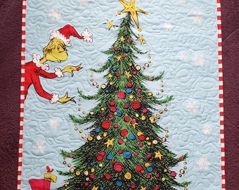 May include: A green Grinch character decorates a Christmas tree with ornaments and a star on top. The tree is surrounded by wrapped presents and a toy bike and train. The background is a blue quilted fabric with snowflakes.