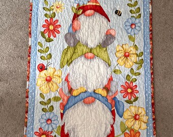 May include: A quilted wall hanging featuring three gnomes stacked on top of each other. The gnomes are wearing red, green, and blue hats and have white beards. The background is a light blue with yellow, red, and blue flowers.