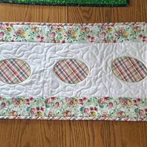 May include: A white quilted table runner with a green floral print border and five oval cutouts with plaid fabric inserts.