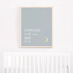 Op de afbeelding: Een ingelijste print met een lichtblauwe achtergrond toont de tekst "i love you to the moon and back" in het wit. Een gele halve maan en sterren staan in de rechter benedenhoek. Het frame is van licht hout. De print hangt boven een witte wieg.