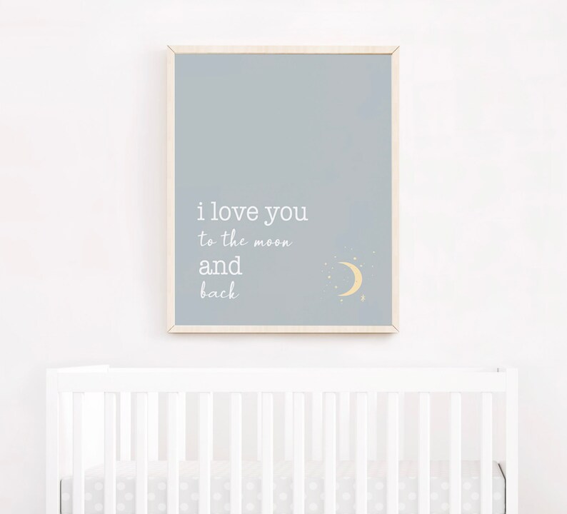 Op de afbeelding: Een ingelijste print met een lichtblauwe achtergrond toont de tekst "i love you to the moon and back" in het wit. Een gele halve maan en sterren staan in de rechter benedenhoek. Het frame is van licht hout. De print hangt boven een witte wieg.