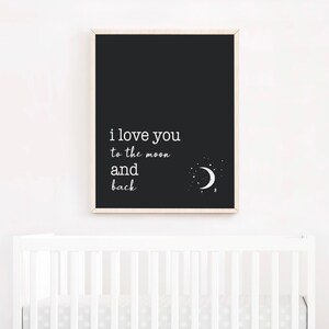 Op de afbeelding: Een zwart-wit print met de tekst "i love you to the moon and back". De print toont een illustratie van een maan en sterren. Het kunstwerk wordt boven een witte wieg getoond.