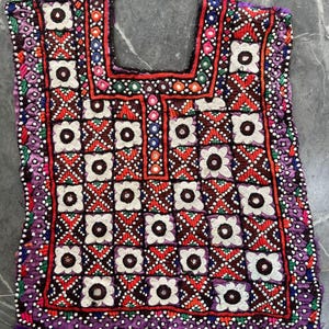 May include: A sleeveless tunic featuring intricate hand-stitched embroidery in a grid pattern. The design incorporates white floral motifs with dark centers, small reflective mirrors, and vibrant red, green, and purple thread accents throughout.