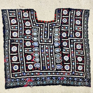 May include: A black textile with intricate embroidery and mirror embellishments. The design features geometric patterns in white, red, and blue, creating a striking visual contrast. The garment has a square shape with a U-shaped neckline, suggesting it is a top or tunic.