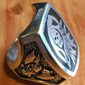 May include: A silver-colored ring with a skull and crossbones design. The ring has a rectangular face with a black background and a detailed skull and skeleton graphic. The band features additional skull and bone details.