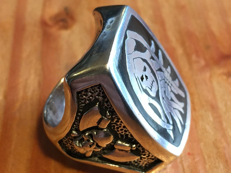 May include: A silver-colored ring with a skull and crossbones design. The ring has a rectangular face with a black background and a detailed skull and skeleton graphic. The band features additional skull and bone details.
