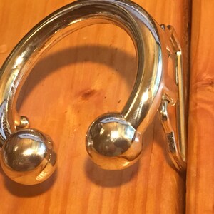May include: A gold-toned metal hook with a rounded, curved design and two ball-shaped ends. The hook is mounted on a wooden surface.