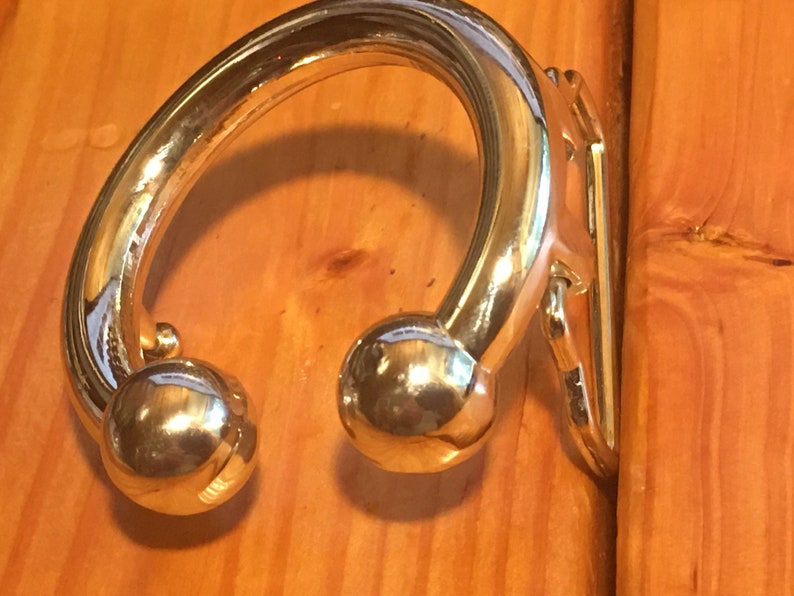 May include: A gold-toned metal hook with a rounded, curved design and two ball-shaped ends. The hook is mounted on a wooden surface.