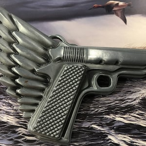 May include: A silver-colored belt buckle shaped like a gun with wings. The gun has a textured grip and a detailed design. The wings are made of multiple feathers.