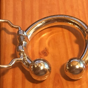 May include: A silver belt buckle with a round, smooth, ring-shaped design. The ring has two round, smooth, silver balls on either side. The buckle has a standard belt loop.