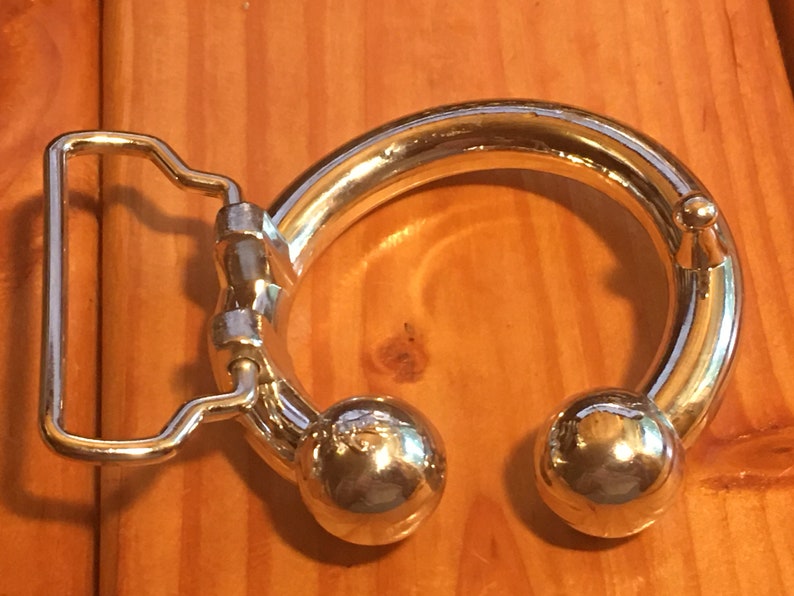 May include: A silver belt buckle with a round, smooth, ring-shaped design. The ring has two round, smooth, silver balls on either side. The buckle has a standard belt loop.