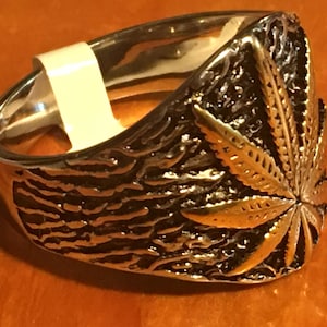 May include: A silver ring with a gold-colored marijuana leaf design. The ring has a textured band.