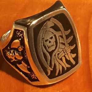 May include: A silver-colored ring with a rectangular face featuring a Grim Reaper design. The ring's face has a black background with a detailed engraving of the Grim Reaper holding a scythe. The sides of the ring feature skull designs.